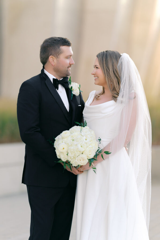 Bride and groom smiling at each other while holding a white rose wedding bouquet